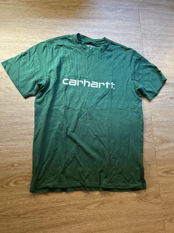 Camiseta Carhartt verde tirada sobre un suelo de parquet, fotografiada con un móvil