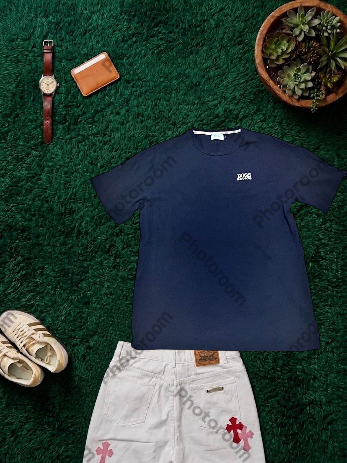 Resultado de un SaaS de fotos: camiseta azul marino sobre césped artificial, con reloj, cartera, planta, zapatillas y un pantalón alrededor, y su marca de agua encima.