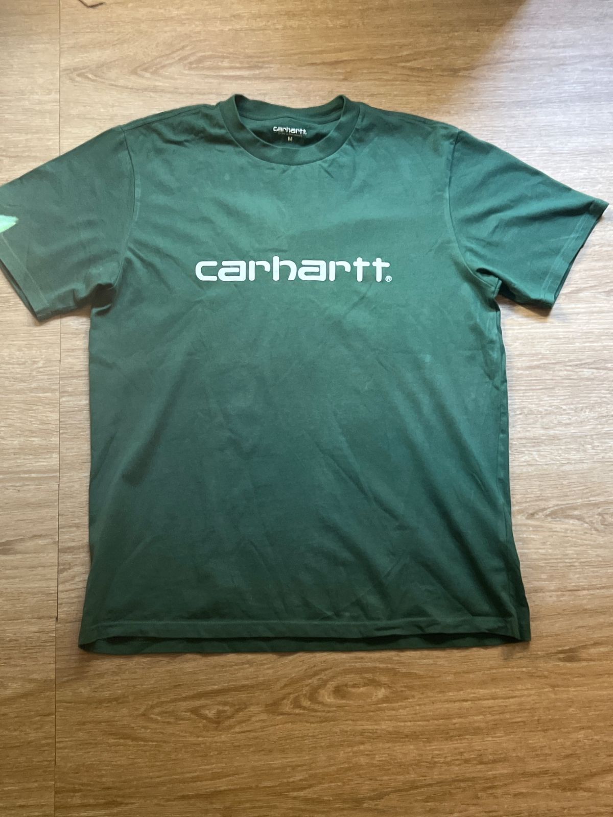 La foto del móvil: una camiseta verde Carhartt sobre el suelo de madera de una casa